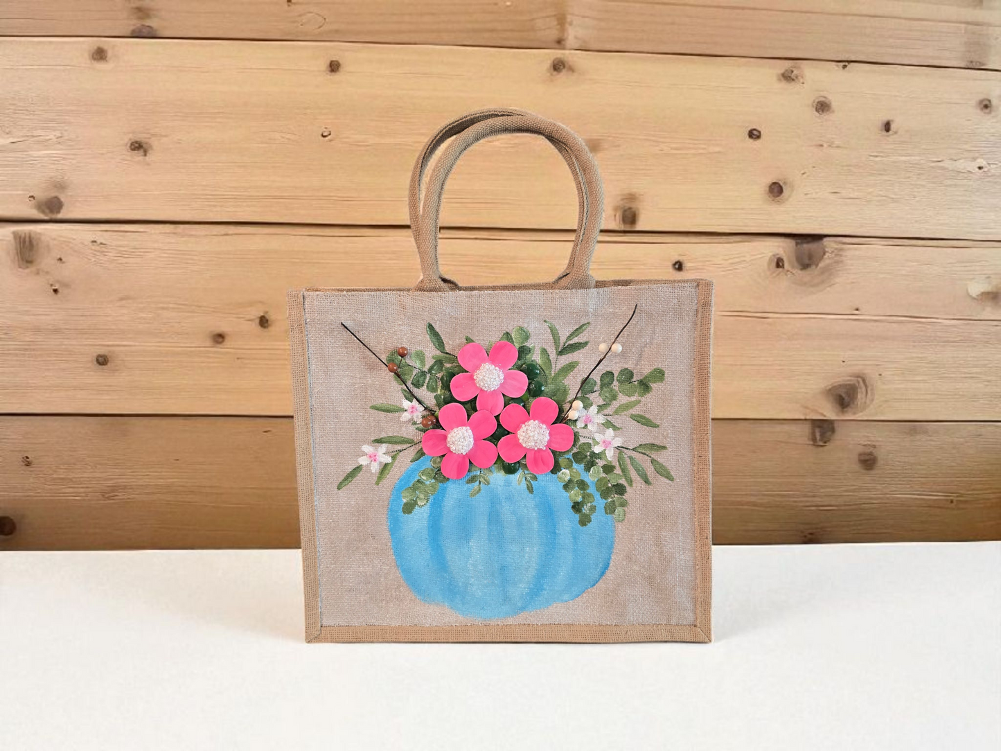 Hand-painted jute tote featuring a blue pumpkin used as a flower pot, filled with pink flowers and greenery; painted on off-white metallic background with beads, leaves, and wooden floral embellishments