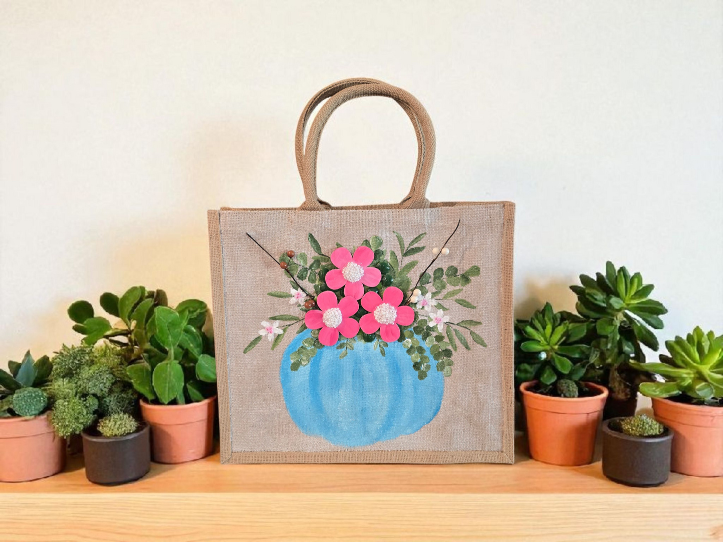 Hand-painted jute tote featuring a blue pumpkin used as a flower pot, filled with pink flowers and greenery; painted on off-white metallic background with beads, leaves, and wooden floral embellishments