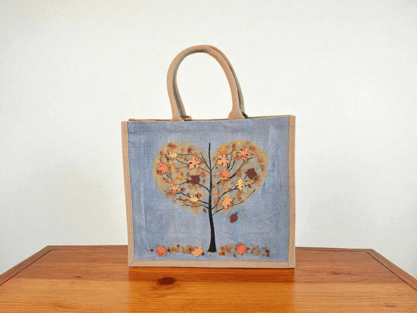 Hand-painted jute tote featuring a heart-shaped fall tree with colorful leaves in red, orange, and gold; painted on a pale blue metallic background with beads and wooden embellishments