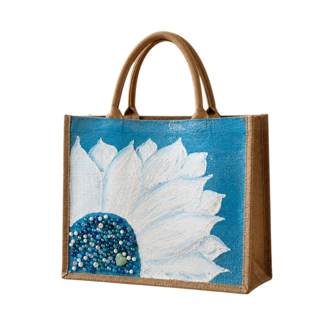 Hand-painted jute tote bag featuring oversized white floral petals on a bright turquoise blue background with blue, pearl, and crystal bead embellishments at the flower center, finished with natural jute trim and rounded padded handles.