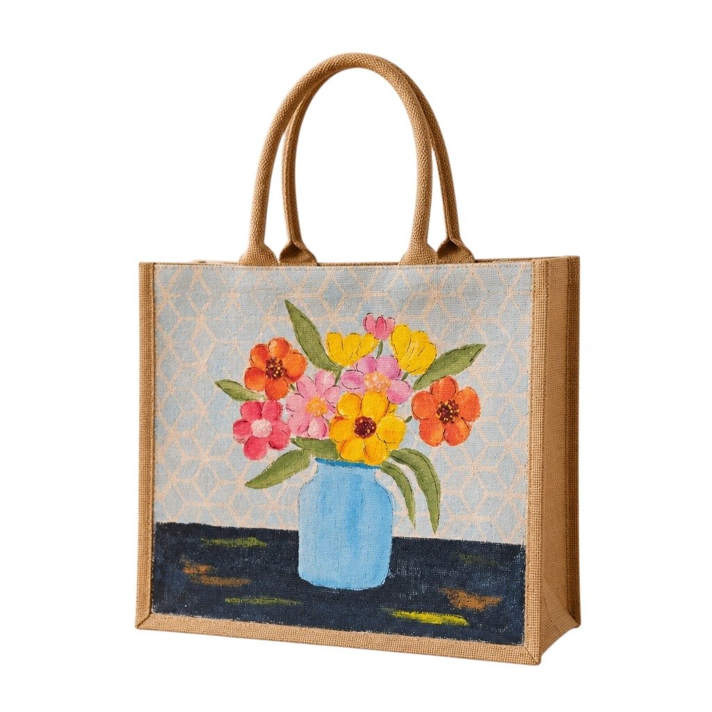 Hand-painted jute tote bag featuring a colorful floral bouquet in a light blue vase against a soft patterned background. The design includes bright pink, yellow, and orange flowers with green leaves, creating a cheerful cottage-inspired look with natural woven handles and artisan charm.