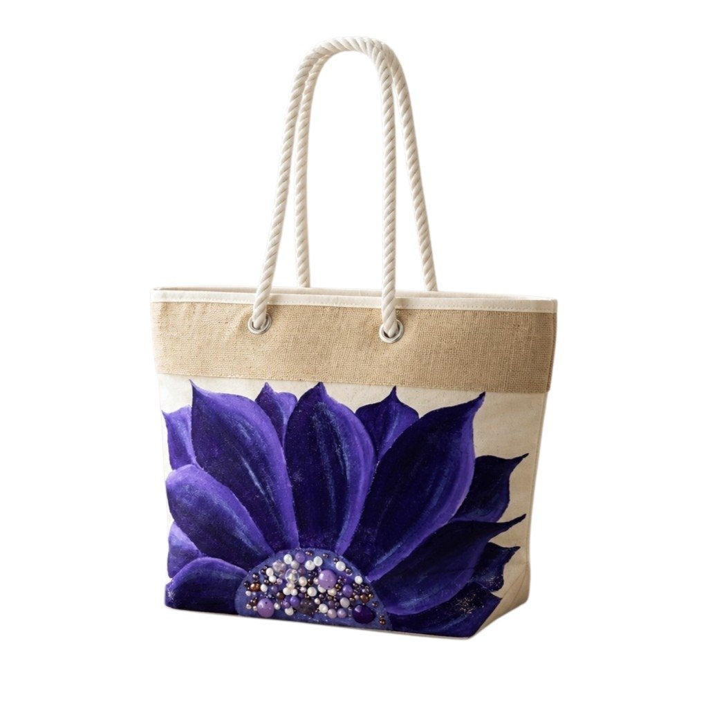 Hand-painted rope handle beach tote featuring a bold purple floral design with jeweled embellishments at the flower center, accented with natural jute trim and oversized cream rope handles against a neutral background.