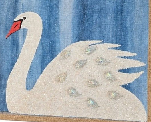 Close up of Hand-painted jute tote bag featuring a white swan with jeweled feather accents on a textured blue background, finished with natural braided handles and coastal-inspired style.