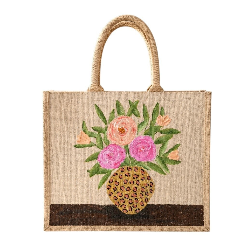 Hand-painted natural jute tote bag featuring a floral arrangement of pink and peach blooms with green leaves in a leopard-print vase. The structured tote has natural woven handles and a warm neutral palette that blends feminine floral charm with trendy animal-print accents.