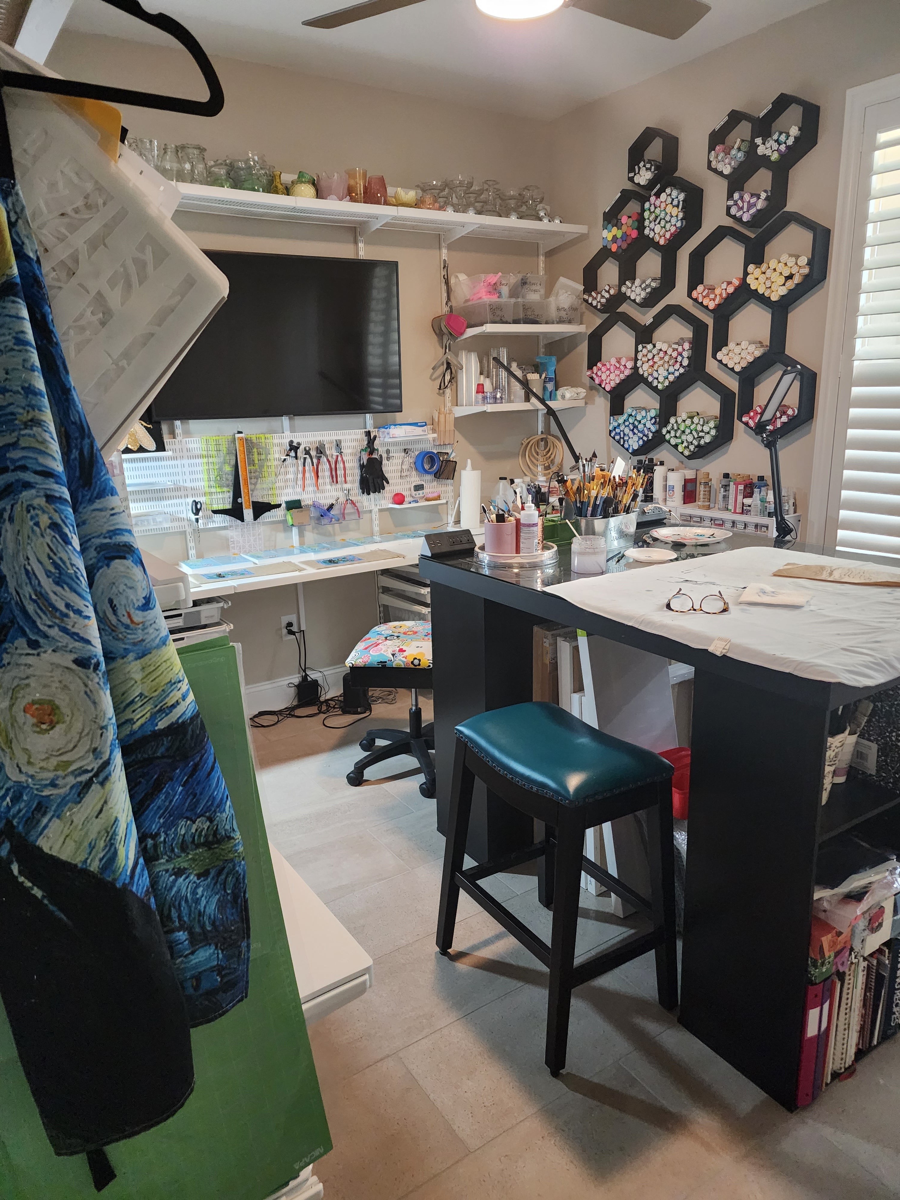 Bright, organized art studio with shelves of glass embellishments, paintbrushes, tools, and a central worktable used for hand-painted tote bag designs.