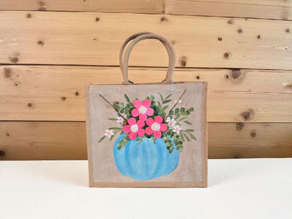 Hand-painted jute tote featuring a blue pumpkin used as a flower pot, filled with pink flowers and greenery; painted on off-white metallic background with beads, leaves, and wooden floral embellishments