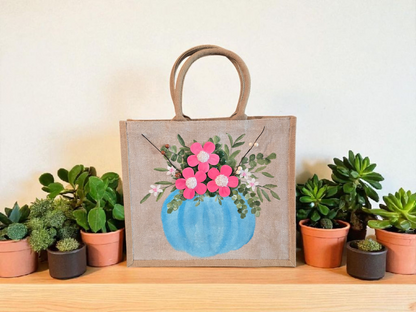 Hand-painted jute tote featuring a blue pumpkin used as a flower pot, filled with pink flowers and greenery; painted on off-white metallic background with beads, leaves, and wooden floral embellishments