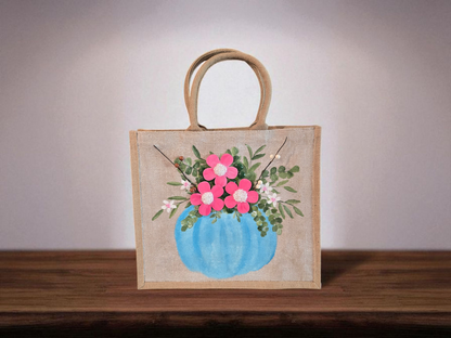 Hand-painted jute tote featuring a blue pumpkin used as a flower pot, filled with pink flowers and greenery; painted on off-white metallic background with beads, leaves, and wooden floral embellishments