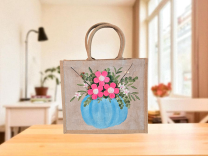 Hand-painted jute tote featuring a blue pumpkin used as a flower pot, filled with pink flowers and greenery; painted on off-white metallic background with beads, leaves, and wooden floral embellishments