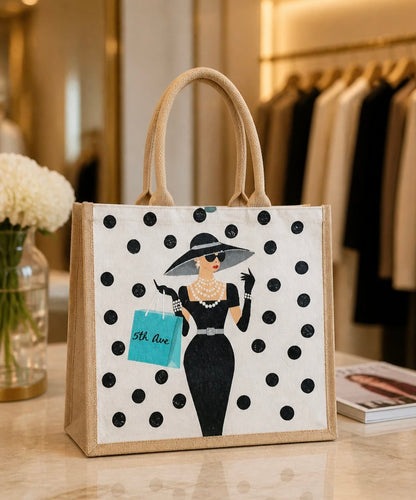 Hand-painted jute tote bag featuring a stylish woman in a black dress and wide-brim hat with pearl jewelry, black polka dot background, and turquoise “5th Ave” shopping bag detail.