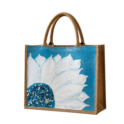 Hand-painted jute tote bag featuring oversized white floral petals on a bright turquoise blue background with blue, pearl, and crystal bead embellishments at the flower center, finished with natural jute trim and rounded padded handles.