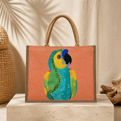 Hand-painted tropical parrot tote bag on natural jute canvas featuring a colorful blue, green, and yellow parrot against a coral-orange background, finished with sturdy double handles and a structured rectangular shape.