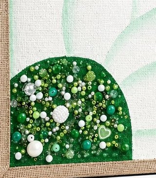 close up of Hand-painted sunflower tote bag featuring large white floral petals on a vibrant green background with sparkling green, pearl, and crystal embellishments at the flower center, accented with natural jute trim and rounded padded handles against a neutral background.