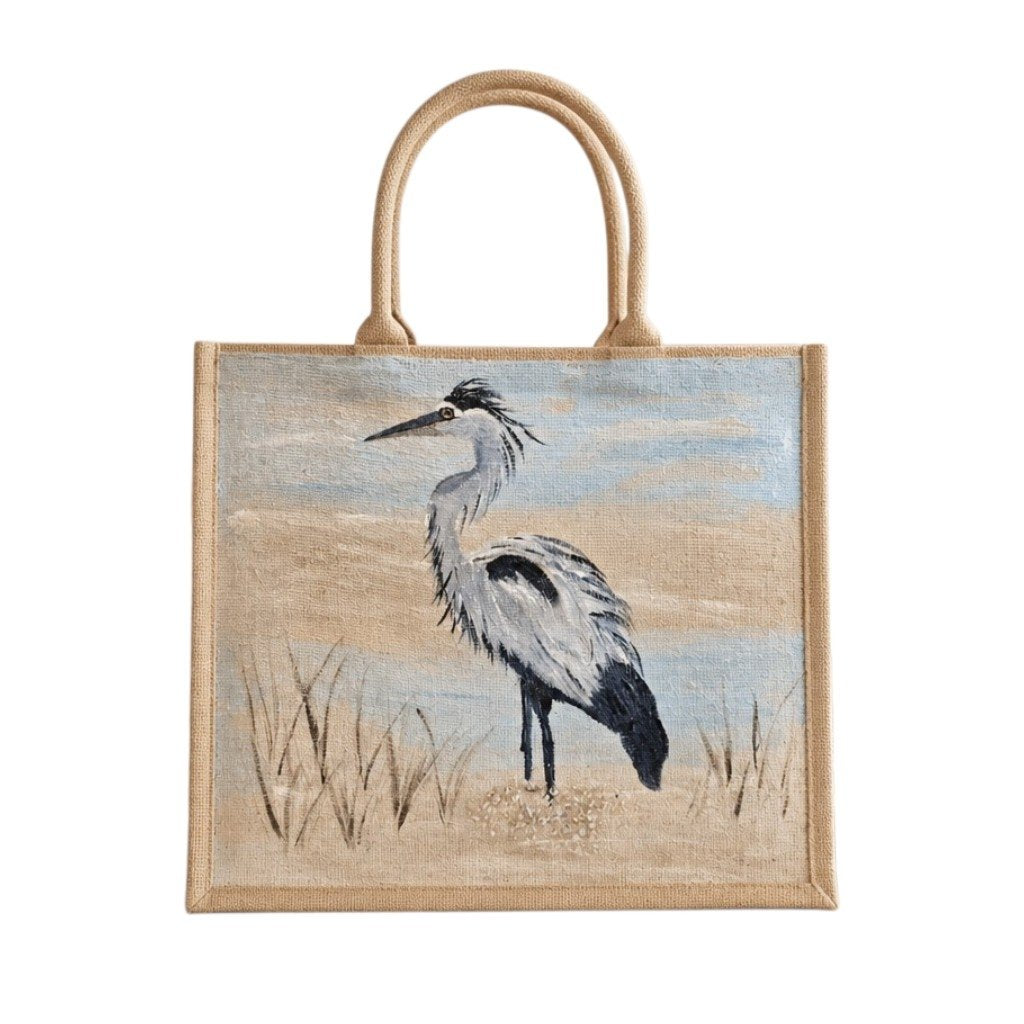 Hand-painted coastal heron tote bag on natural jute canvas featuring a graceful gray and white heron standing in marsh grass against a soft blue and sandy background.