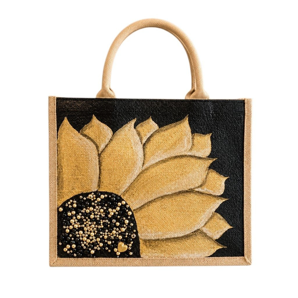 Hand-painted sunflower tote bag featuring soft gold petals on a black background with pearl and gold jeweled embellishments at the flower center, accented with natural jute trim and rounded padded handles against a neutral background.