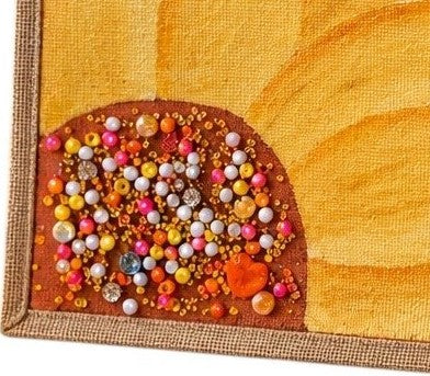 Close up of Hand-painted sunflower tote bag featuring soft yellow floral petals on a warm orange background with colorful jeweled embellishments at the flower center, accented with natural jute trim and rounded padded handles against a neutral background.