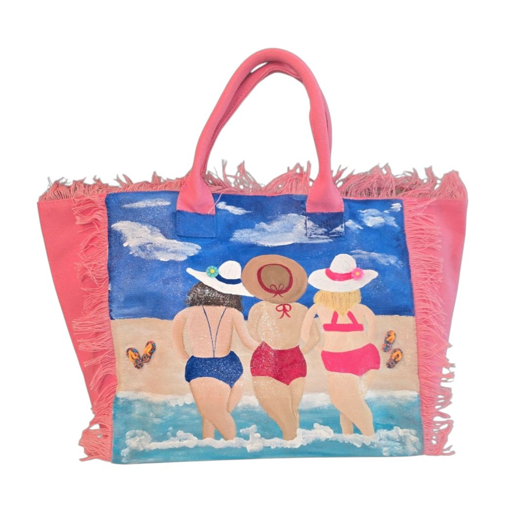 Hand-painted pink fringed tote bag featuring three women walking into the ocean in colorful swimsuits and sun hats, with a beach scene, blue sky, clouds, and flip-flop details on a vibrant coastal-inspired design.