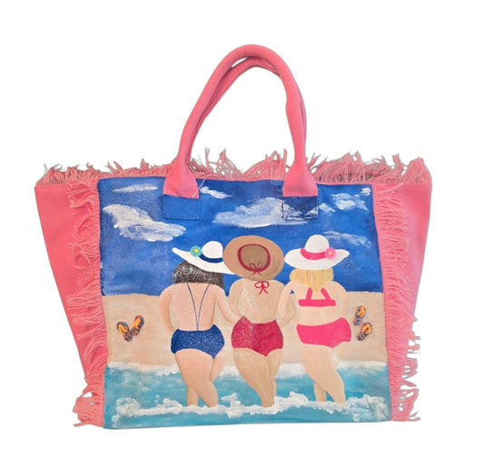 Hand-painted pink fringed tote bag featuring three women walking into the ocean in colorful swimsuits and sun hats, with a beach scene, blue sky, clouds, and flip-flop details on a vibrant coastal-inspired design.