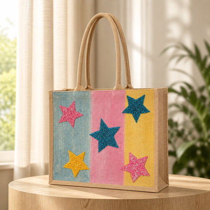 Colorful hand-painted jute tote bag featuring pastel blue, pink, and yellow panels accented with glittery star designs in teal, pink, and gold. The structured tote has natural woven handles and a bright, playful aesthetic inspired by fun artisan fashion accessories.