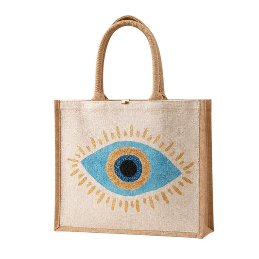 Hand-painted jute tote bag featuring a blue and gold evil eye design centered on a natural canvas background, accented with metallic gold detailing, natural jute trim, and rounded padded handles.