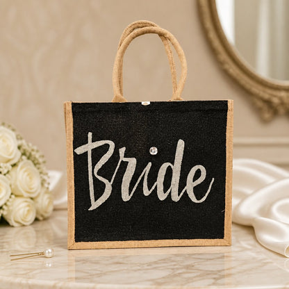 Hand-painted black jute tote bag featuring the word “Bride” in bold white script with a rhinestone accent, designed for weddings, bridal celebrations, and stylish bride-to-be gifts.
