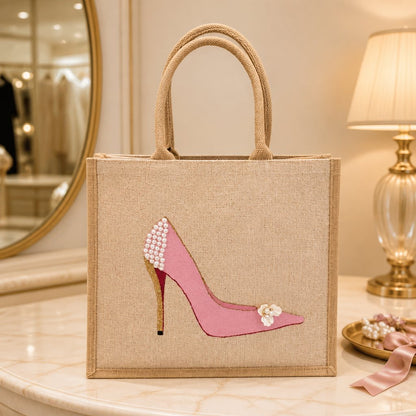 Hand-painted pink stiletto tote bag on natural jute canvas featuring pearl embellishments on the heel, a gold glitter heel accent, and delicate floral detail on the toe.