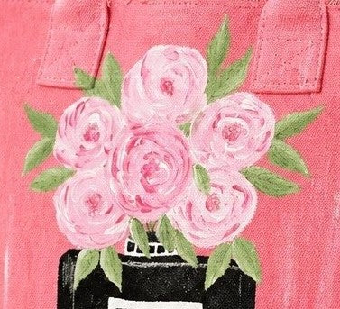 close up of Pink fringed canvas tote bag featuring a hand-painted black perfume bottle vase filled with soft pink floral blooms and green leaves. The bag has textured fringe detailing along the edges, matching pink handles, and a feminine boutique-inspired design with playful artisan charm.
