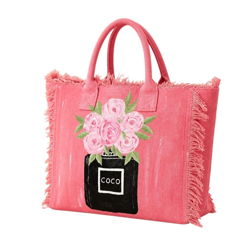 Pink fringed canvas tote bag featuring a hand-painted black perfume bottle vase filled with soft pink floral blooms and green leaves. The bag has textured fringe detailing along the edges, matching pink handles, and a feminine boutique-inspired design with playful artisan charm.
