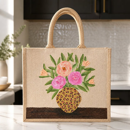 Hand-painted natural jute tote bag featuring a floral arrangement of pink and peach blooms with green leaves in a leopard-print vase. The structured tote has natural woven handles and a warm neutral palette that blends feminine floral charm with trendy animal-print accents.