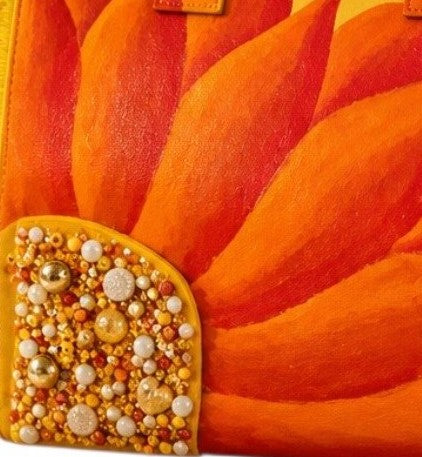 close up of Hand-painted yellow fringed tote bag featuring a bold sunflower design in vibrant orange and red tones, accented with pearl and gold embellishments on the flower center, displayed against a neutral background.
