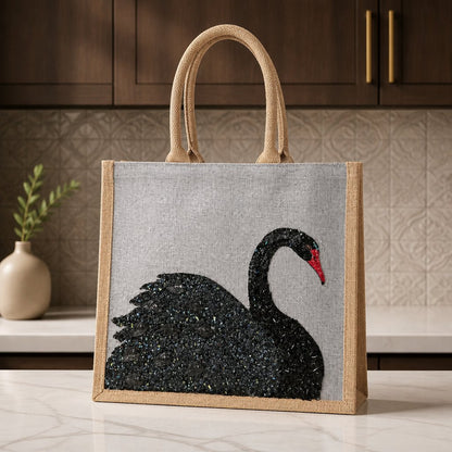 Hand-painted gray jute tote bag featuring an embellished black swan design with textured shimmer details and a red beak. The structured tote has natural woven handles and a sophisticated neutral color palette inspired by elegant, artisan-style fashion accessories.
