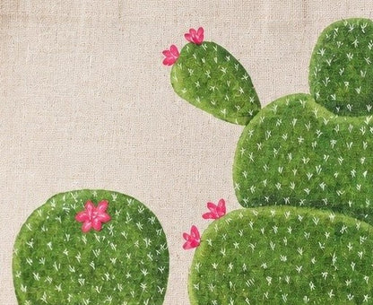 Close up of Hand-painted cactus tote bag featuring whimsical green cactus designs with pink blooms on a natural canvas background, accented with jute trim and rounded padded handles against a neutral background.