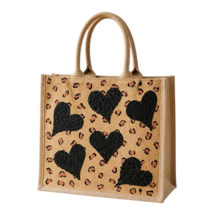 Hand-painted jute shopper bag featuring textured black hearts over a tan background with scattered leopard print accents and natural tan handles.