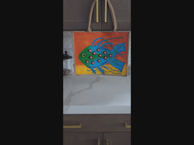 Video of Hand-painted jute tote bag featuring a whimsical blue and green fish with dimensional coral-colored accents on a vibrant orange and yellow background. Coastal-inspired artisan beach tote with rope handles and colorful tropical ocean design.