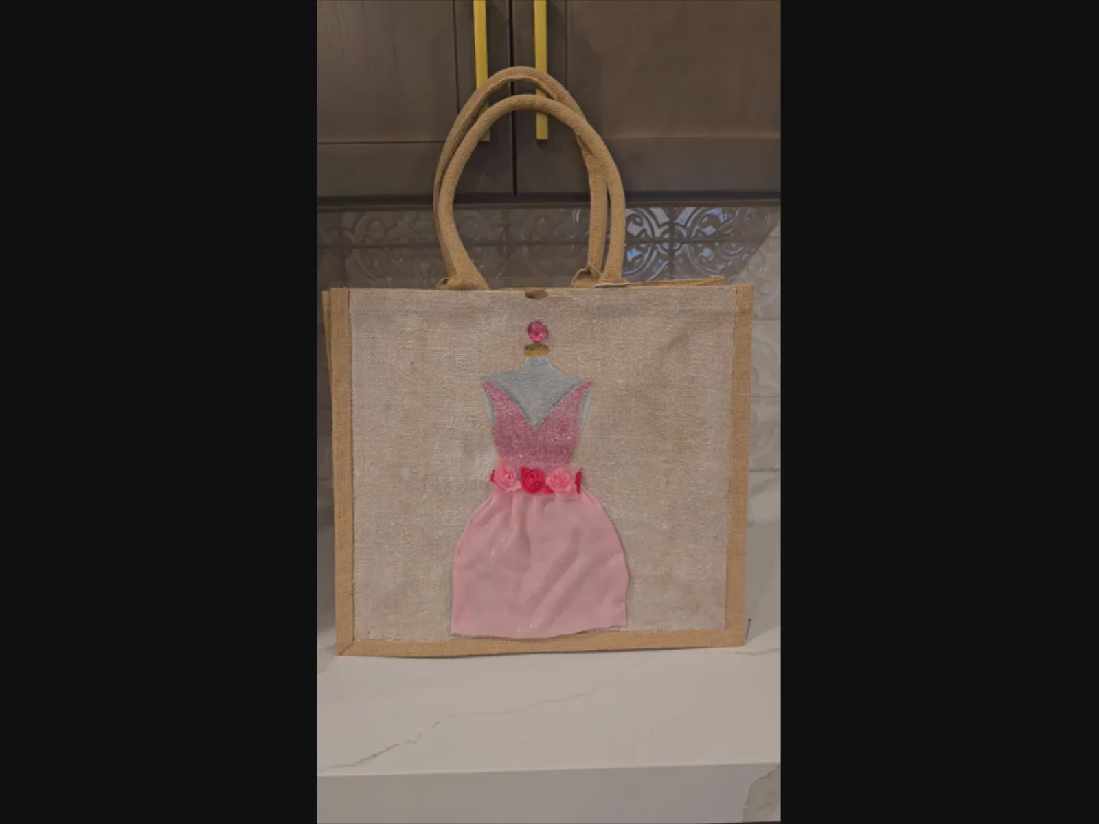 video ofHand-painted jute tote bag featuring a pink embellished cocktail dress design with floral waist details, rhinestone accents, and layered textured skirt on a natural canvas background. 