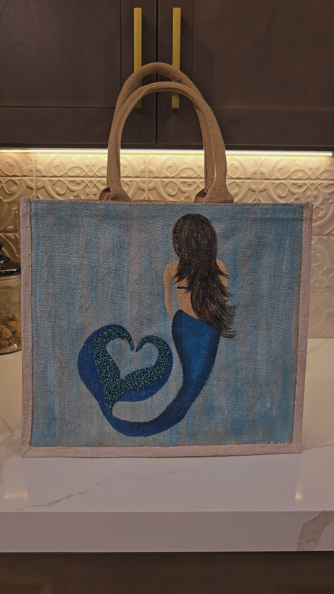 Video of Hand-painted jute tote bag featuring a blue mermaid with flowing brown hair and a heart-shaped tail design on a soft blue background, accented with glitter details and natural burlap handles.