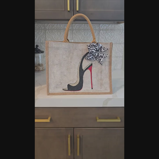 Hand-painted jute tote bag featuring a black stiletto heel with red sole and a zebra-print bow, designed by Dana’s Uncommon Glass