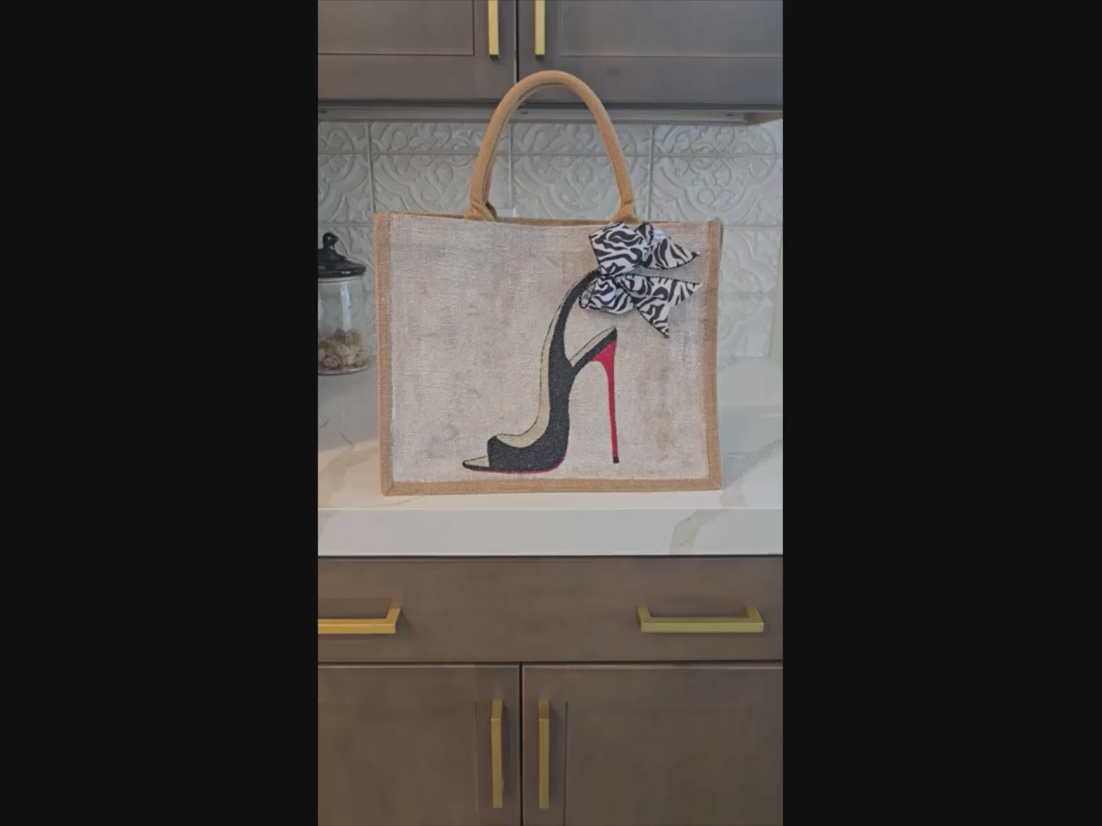 Hand-painted jute tote bag featuring a black stiletto heel with red sole and a zebra-print bow, designed by Dana’s Uncommon Glass