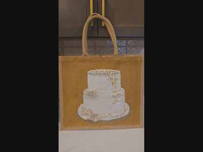 video of Hand-painted jute tote bag featuring an elegant white wedding cake with pearl and floral details on a natural canvas background