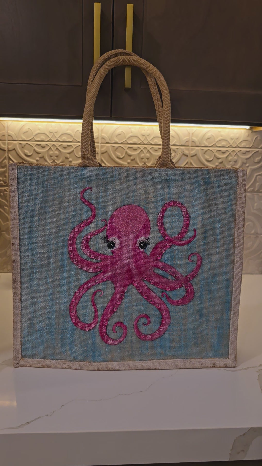 Video of Hand-painted jute tote bag featuring a whimsical pink octopus with curled tentacles and long eyelashes on a soft aqua blue background, finished with natural burlap handles and a beach-inspired coastal style.