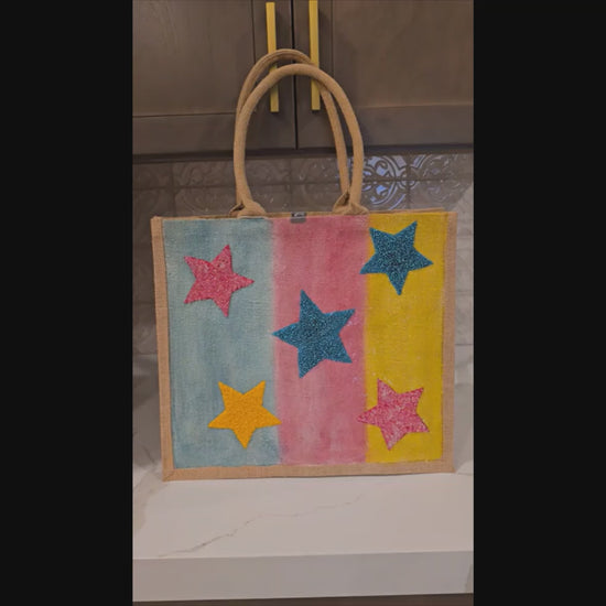 Video of Hand-painted jute tote bag with colorful vertical stripes in blue, pink, and yellow, featuring textured glitter stars in bright colors