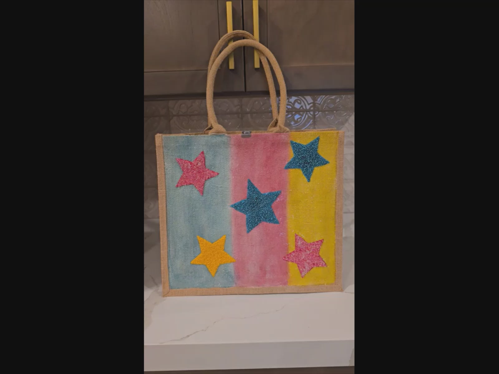 Video of Hand-painted jute tote bag with colorful vertical stripes in blue, pink, and yellow, featuring textured glitter stars in bright colors