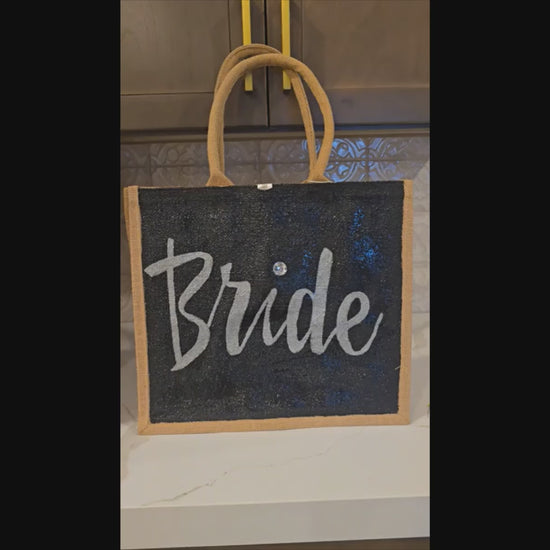 Hand-painted black jute tote bag featuring the word “Bride” in bold white script with a rhinestone accent, designed for weddings, bridal celebrations, and stylish bride-to-be gifts.
