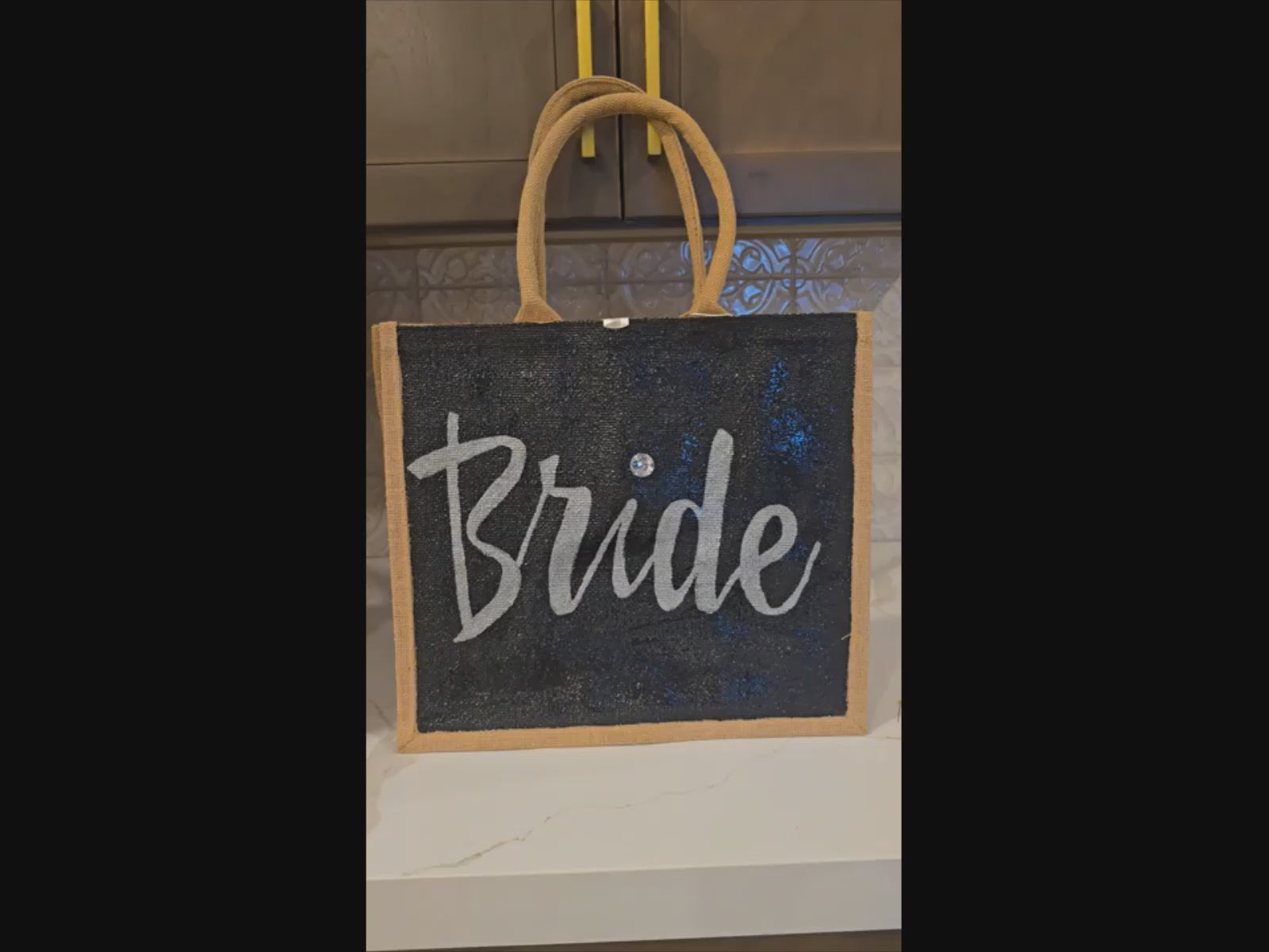 Hand-painted black jute tote bag featuring the word “Bride” in bold white script with a rhinestone accent, designed for weddings, bridal celebrations, and stylish bride-to-be gifts.