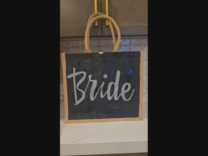 Hand-painted black jute tote bag featuring the word “Bride” in bold white script with a rhinestone accent, designed for weddings, bridal celebrations, and stylish bride-to-be gifts.