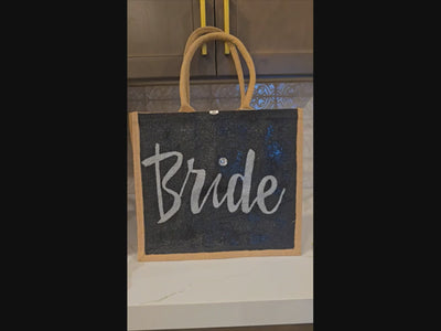 Hand-painted black jute tote bag featuring the word “Bride” in bold white script with a rhinestone accent, designed for weddings, bridal celebrations, and stylish bride-to-be gifts.