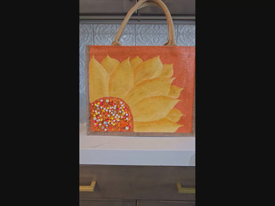Video of Hand-painted sunflower tote bag featuring soft yellow floral petals on a warm orange background with colorful jeweled embellishments at the flower center, accented with natural jute trim and rounded padded handles against a neutral background.
