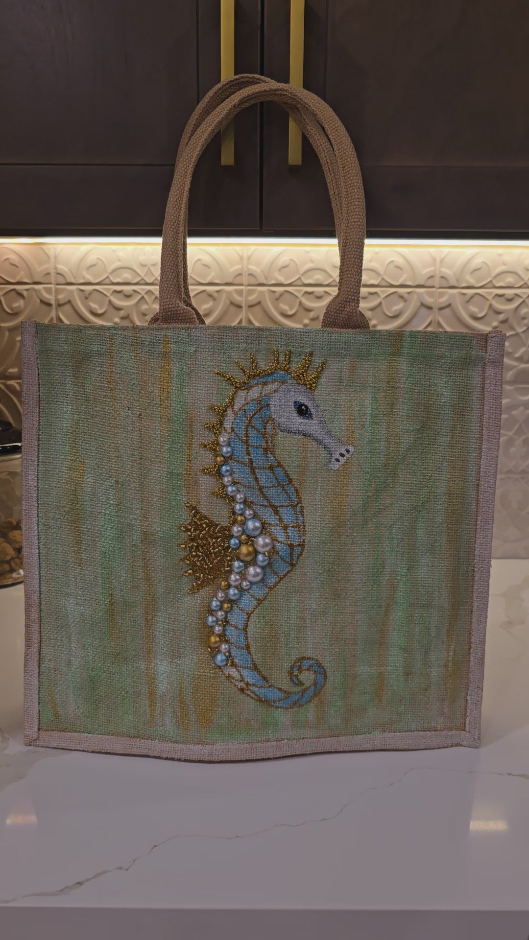 Video of Hand-painted jute tote bag featuring a soft blue seahorse embellished with pearls and gold accents on a textured seafoam green background, finished with natural burlap handles and trim.