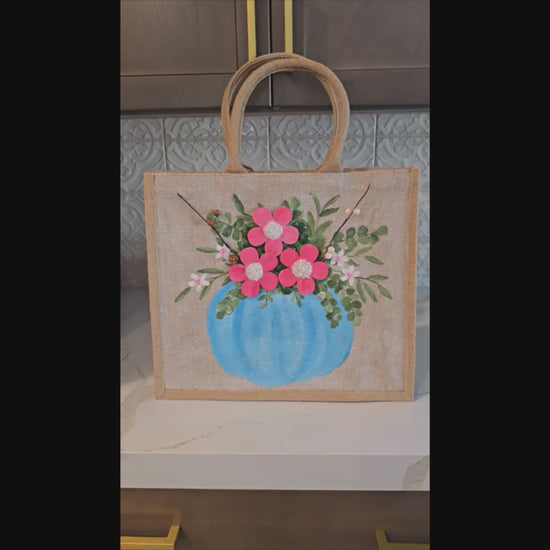 Hand-painted jute tote featuring a blue pumpkin used as a flower pot, filled with pink flowers and greenery; painted on off-white metallic background with beads, leaves, and wooden floral embellishments
