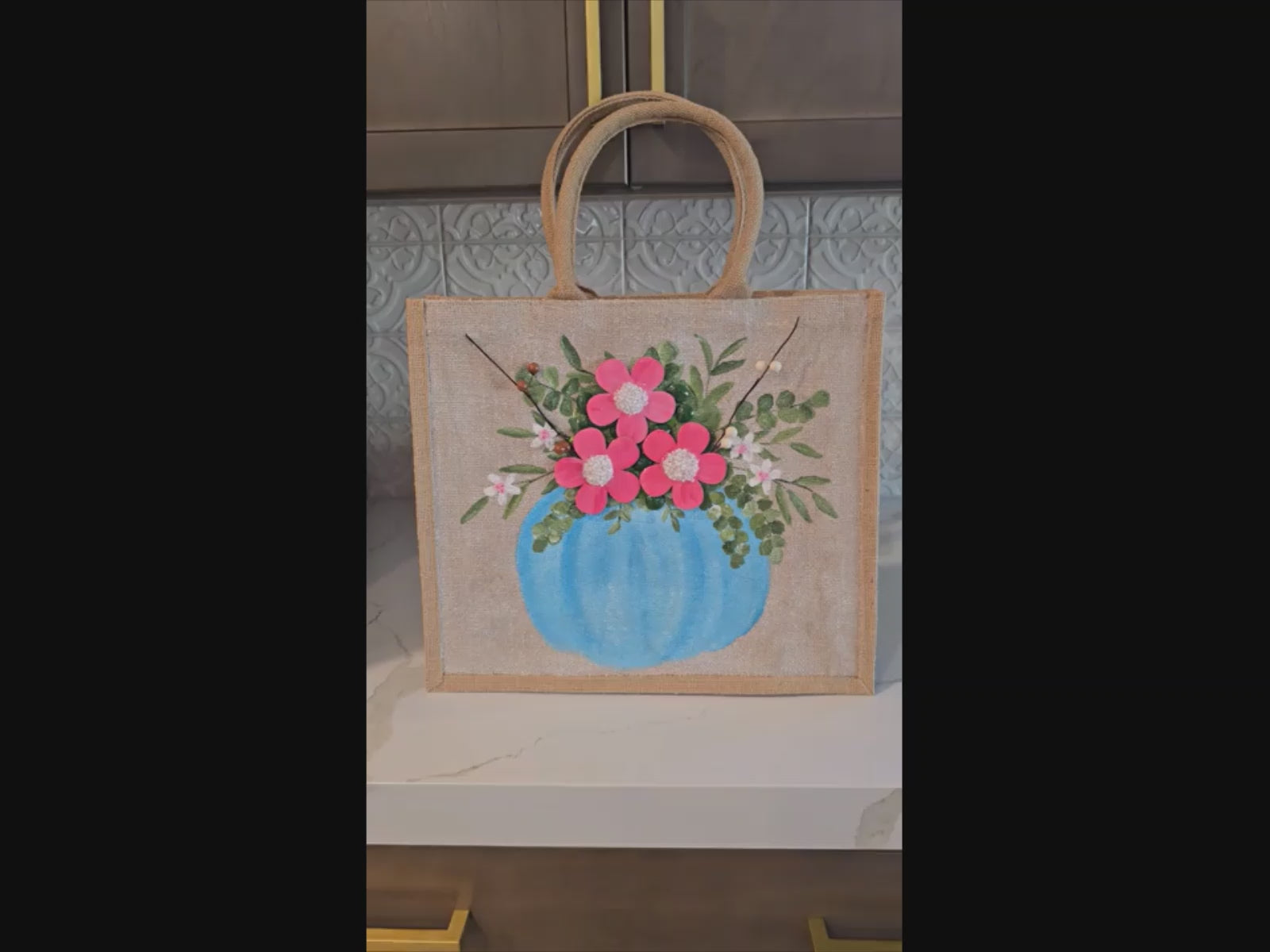 Hand-painted jute tote featuring a blue pumpkin used as a flower pot, filled with pink flowers and greenery; painted on off-white metallic background with beads, leaves, and wooden floral embellishments